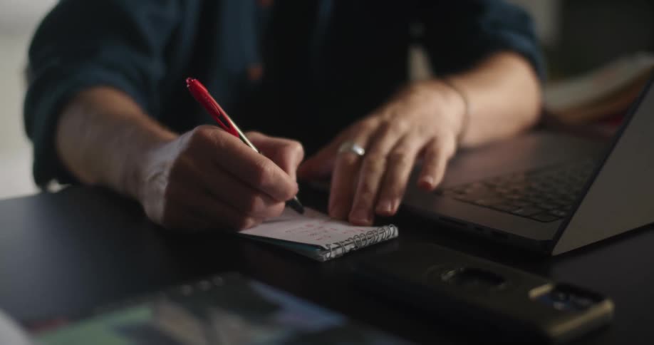 A close up of a man writing with a pen on a notepad next to a laptop
