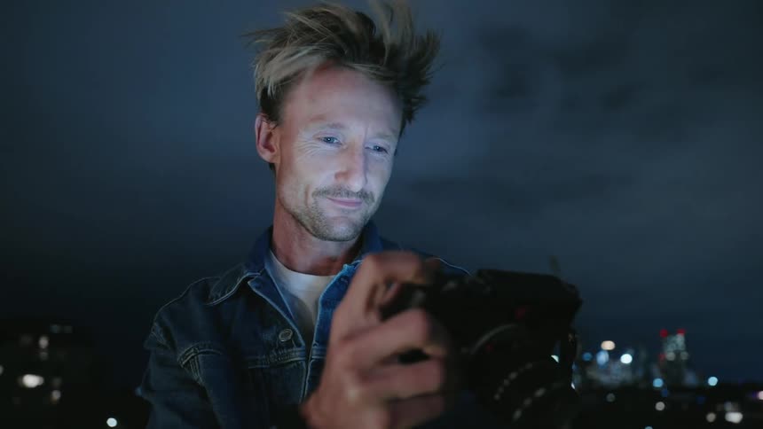Photographer smiles while reviewing photographs as he stands with cityscape in background at night