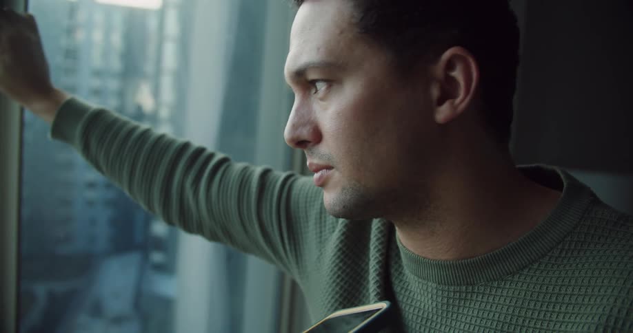 A close up of a man leaning against a window holding his cell phone while talking on it