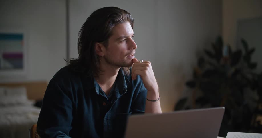 Man sitting at desk working on laptop alternately gazing out window in deep thought
