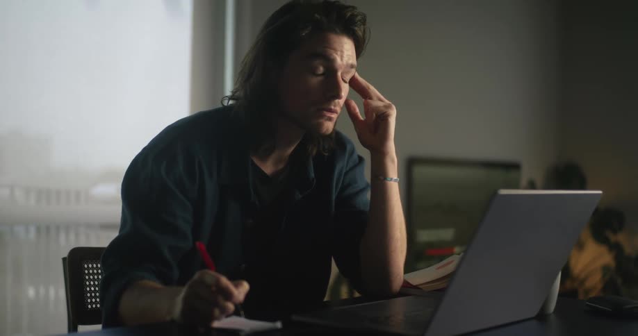 A man is sitting at a table looking at a laptop computer and writing on paper as he appears to be getting tired