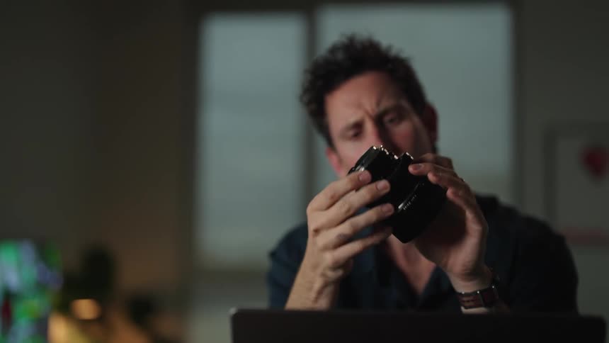 A man holding up a camera lens looking through it as he sits at a desk