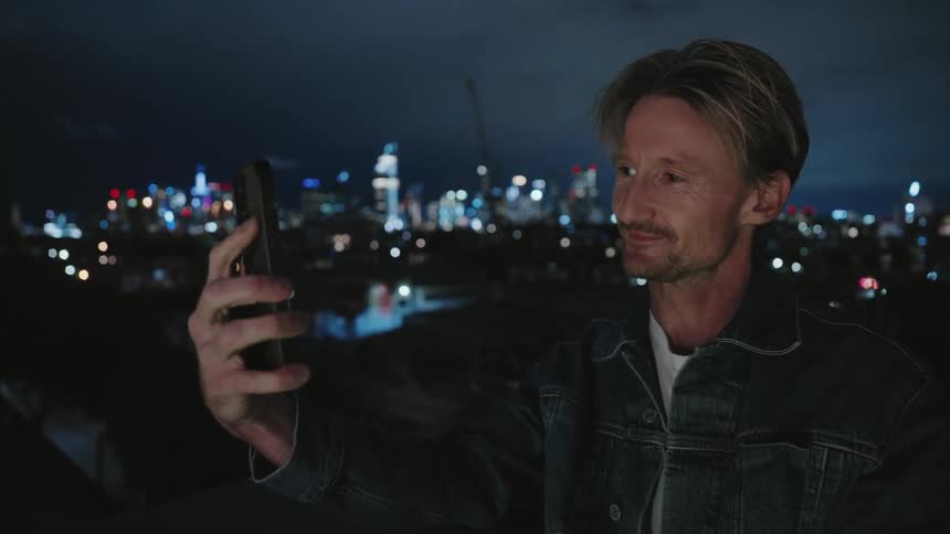 Man waves at screen to end video call on smartphone before standing smiling with night cityscape in the background