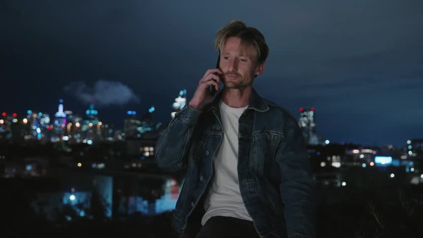 A man sitting outdoors using his cellphone and talking to someone with a city skyline in the background