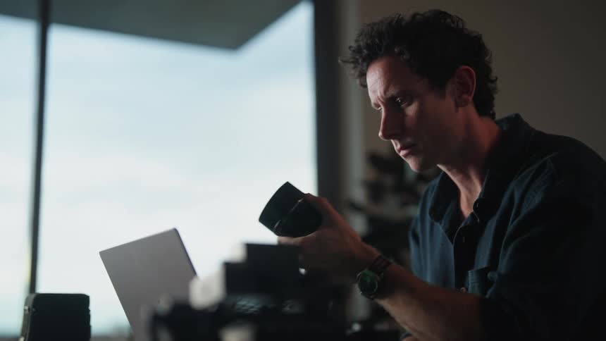 Man sitting at desk in front of laptop and dslr camera examining camera lens