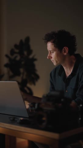 Man sitting at desk with laptop and dslr camera alternately examining camera lens and typing on laptop