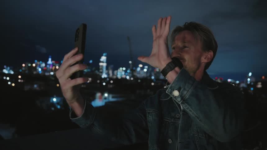 A man standing outside holding his phone up blowing kisses at it and smiling