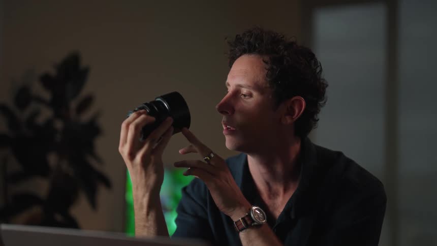 Man sitting at desk with laptop communicating on zoom while handling a camera lens
