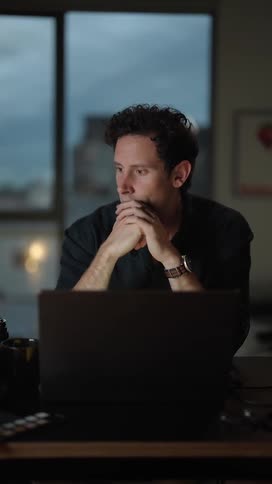 A man is sitting at a table typing on a laptop computer and he pauses to think before continuing