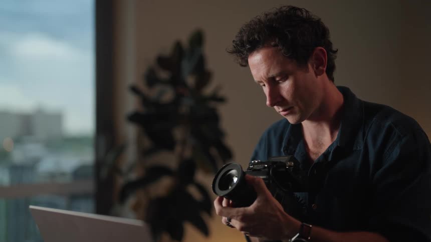A man sitting in front of a laptop picking up a lens and putting it in a camera