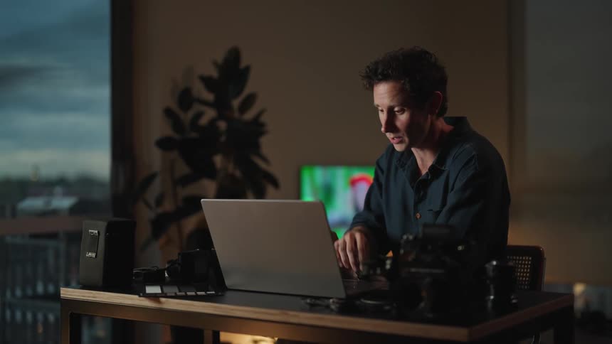 A man is sitting at a table looking at a laptop computer and holding a camera lens in his hands as he has a video conversation and waves to the screen