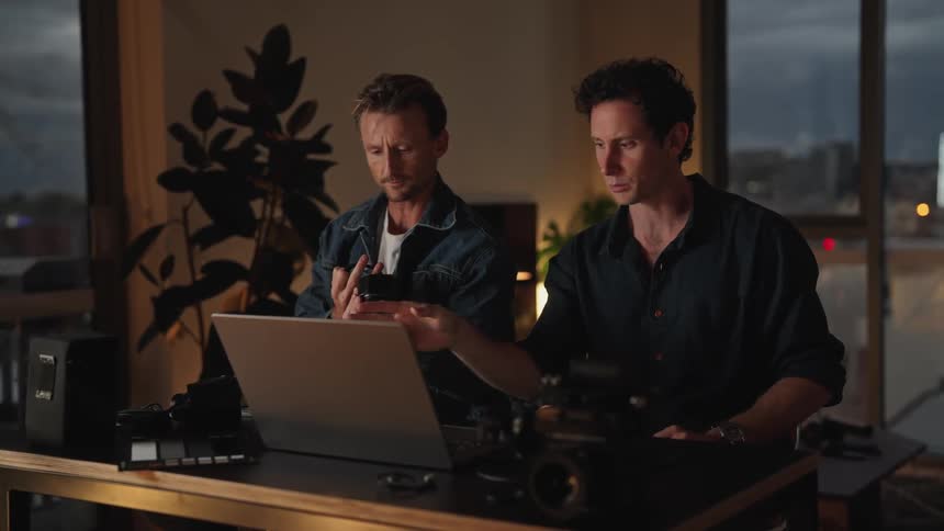 Two male photographers sitting at a desk in front of a laptop discussing and handling cameras and camera equipment