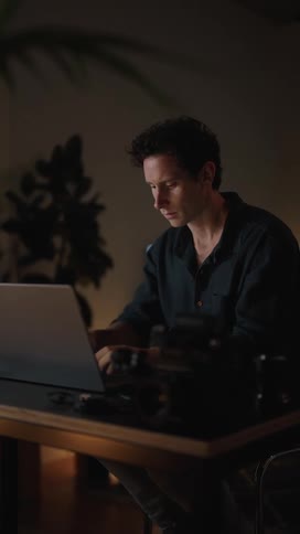 A man is sitting at a table typing on a laptop computer and looking at a camera lens