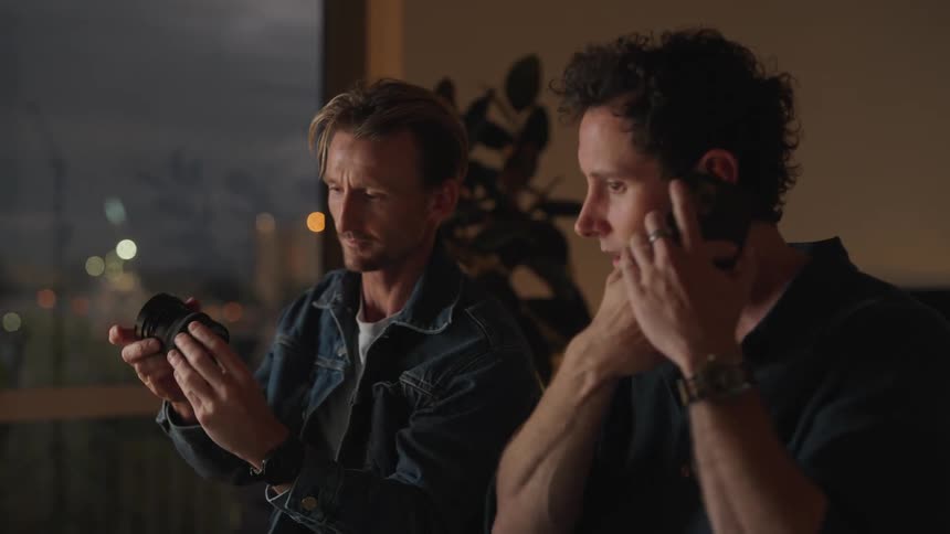 Two men sitting next to each other one on the phone while the other holds up a camera lens to him