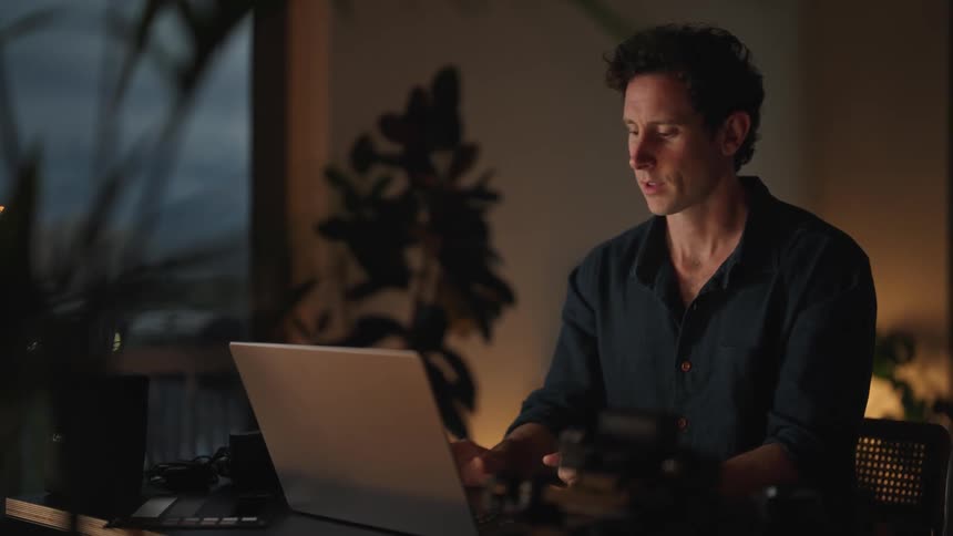 A man sitting at his desk typing on a keyboard and then closing it stretching and getting up