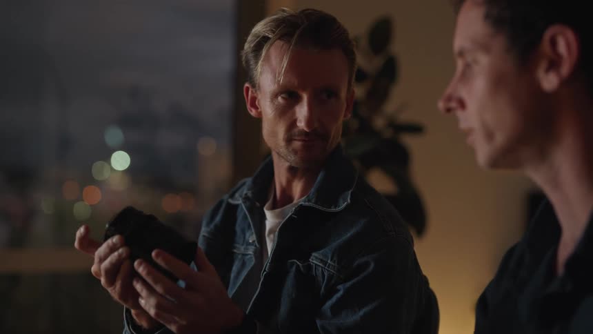 Two men sitting next to each other talking while one holds a camera lens and hands it to the other