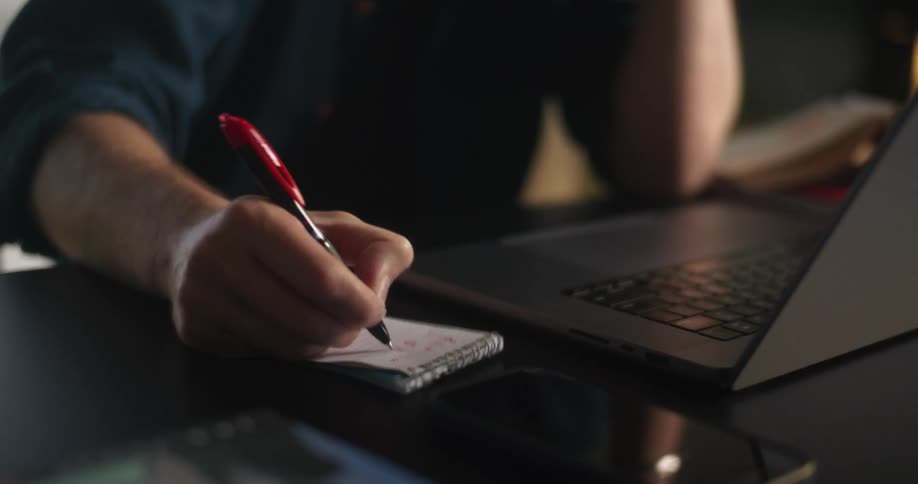 A close up of a perosn writing with a pen on a notepad next to a laptop