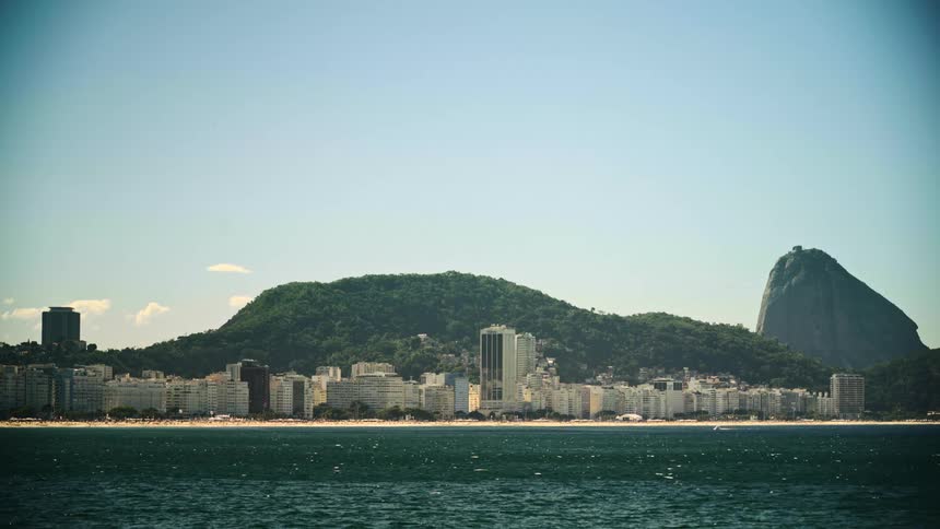 Tall buildings line the coast of copacabana as sunlight reflects on the water and people relax on the beach with a mountain in the distance
