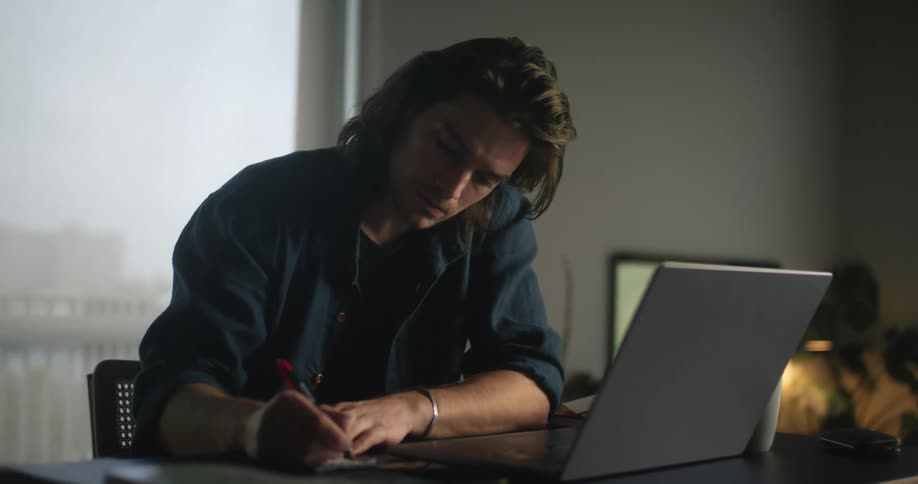 A man is sitting at his computer desk and writing stuff down