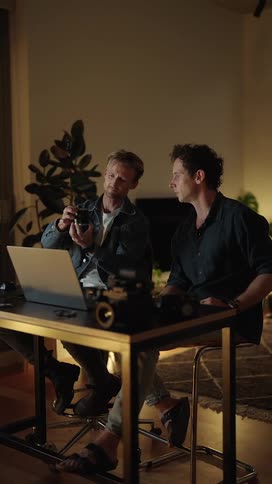 Two men sitting at a desk talking and looking over a camera with a laptop in front of them