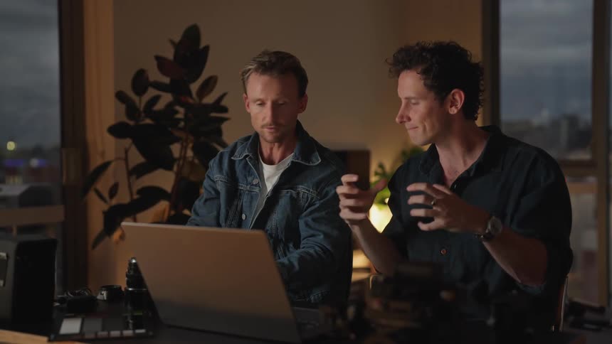 Two men gesture while talking in front of a laptop on a home office at night