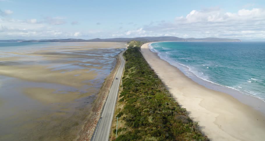 An aerial shot of a beach coastline and a road with a car traveling on it along the coastline