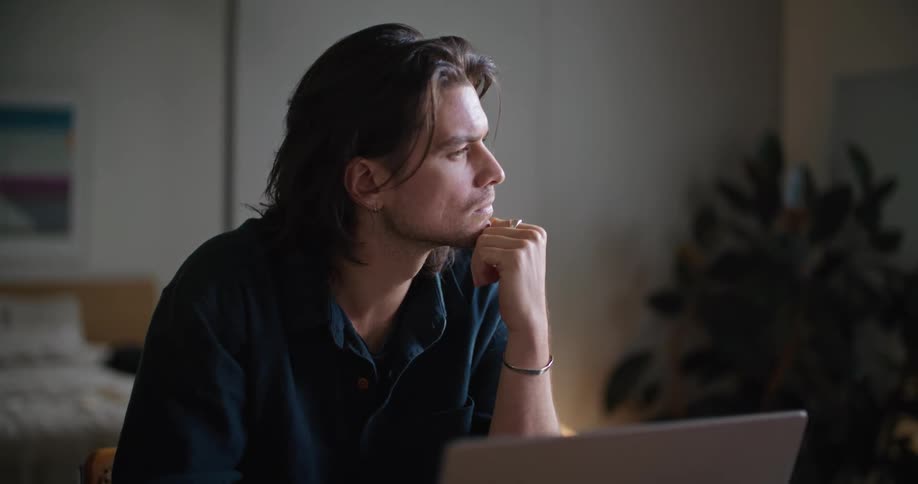 A man sitting at a table in front of a laptop looking off to the left thinking