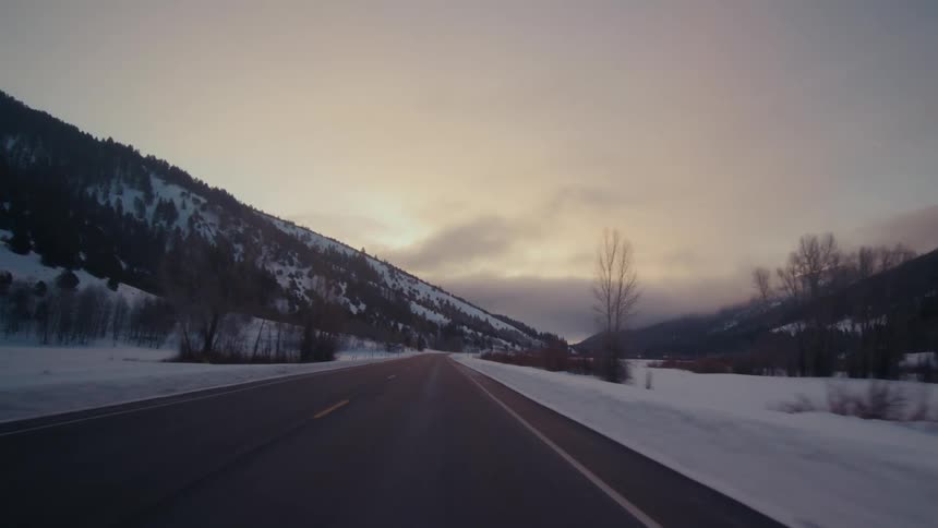 Traveling on a rural road on a snow covered winter evening with forested mountains in the distance under a cloudy sky