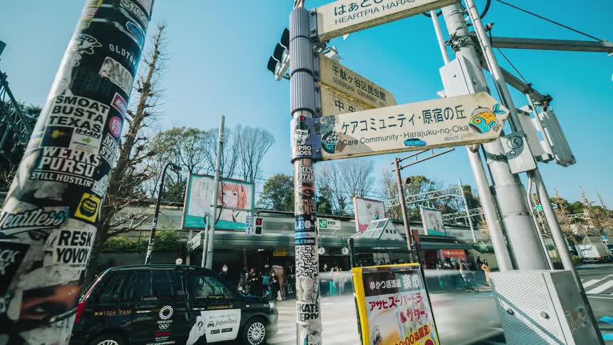 Signs are on poles as traffic moves on the busy street and people walk on the sidewalks as a train travels in the distance