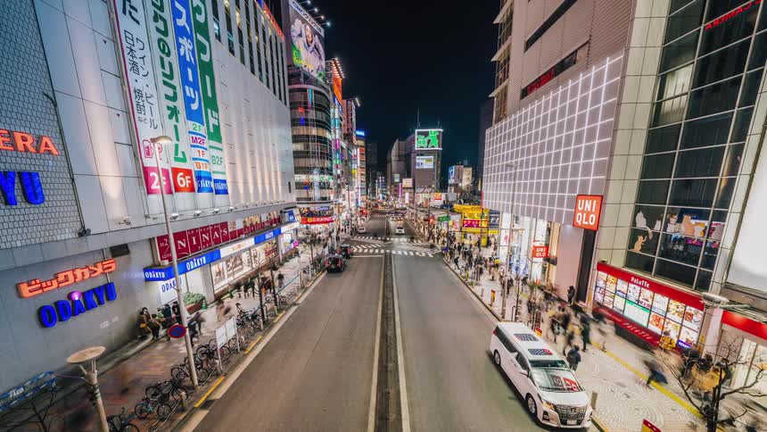 Traffic moves in and out of the busy city as lights glow on tall buildings and people walk on the sidewalks under the night sky