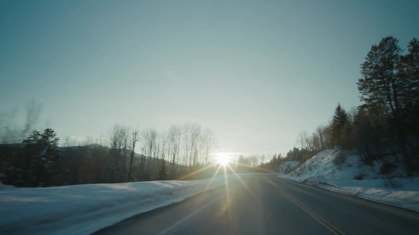 Sun glares from skyover mountains onto highway passing snow covered woods