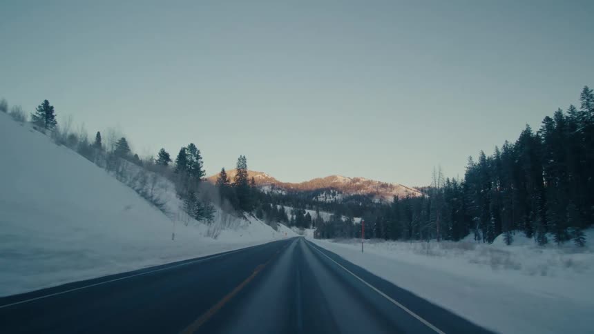 A road traveling through snow covered lands with forests on either side and mountain ranges in the distance