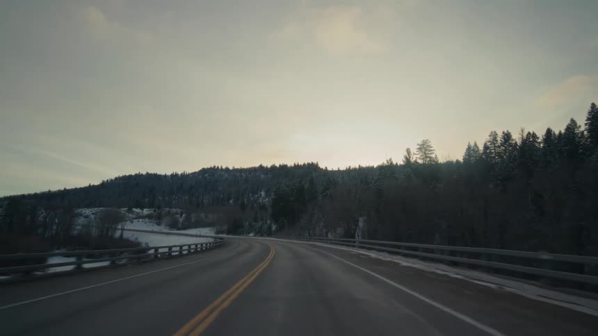 Traveling on a rural highway with snow and trees along the roadside with a bridge over a stream and forested mountains in the distance