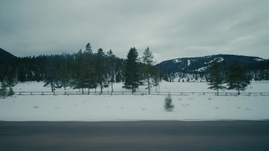Car passes along highway near snow covered pasture and forested mountains