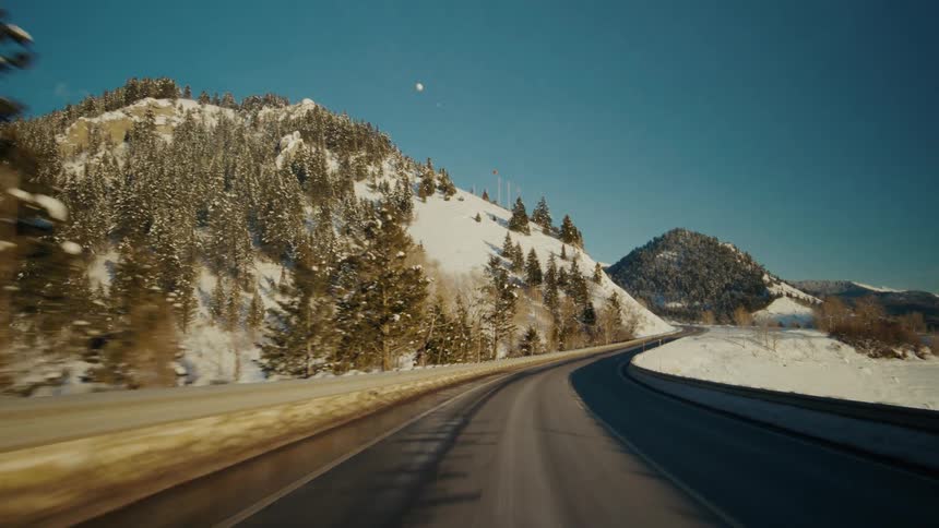 Traveling on a rural highway with snow covered mountains and trees on the roadside and forested mountains in the distance under a clear sky