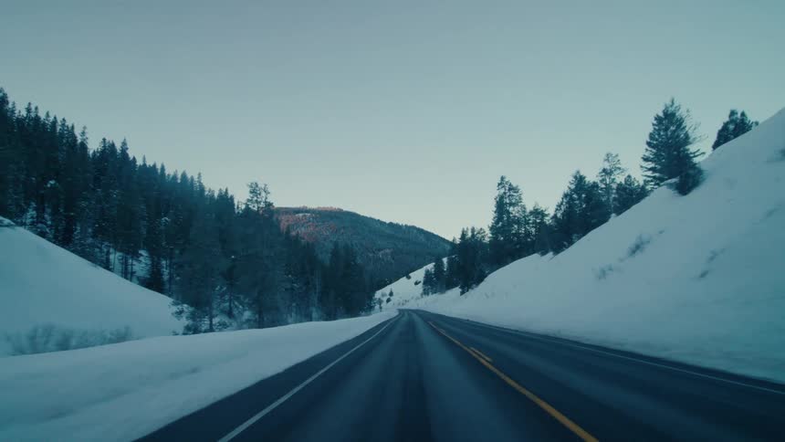 Traveling on a rural highway with snow and trees along the roadside and forested mountains in the distance