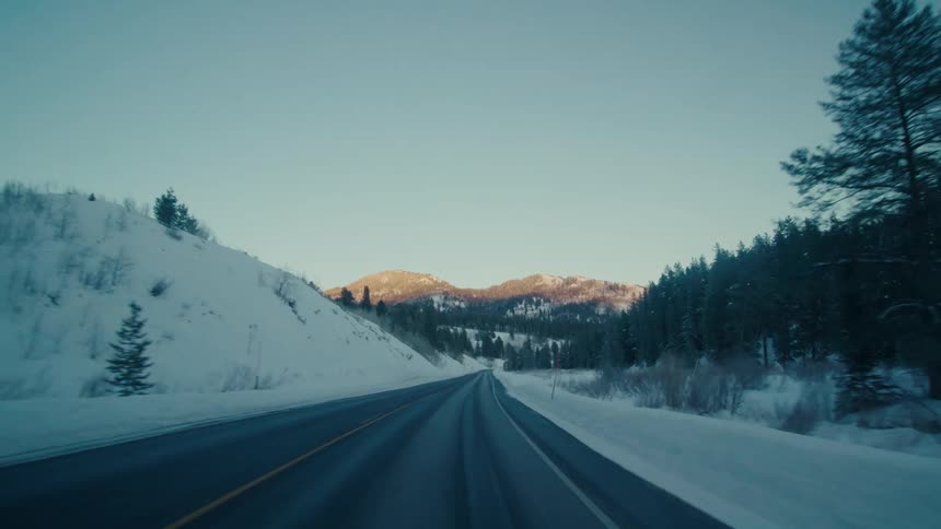 Traveling on a rural highway with snow and trees along the roadside and forested mountains in the distance