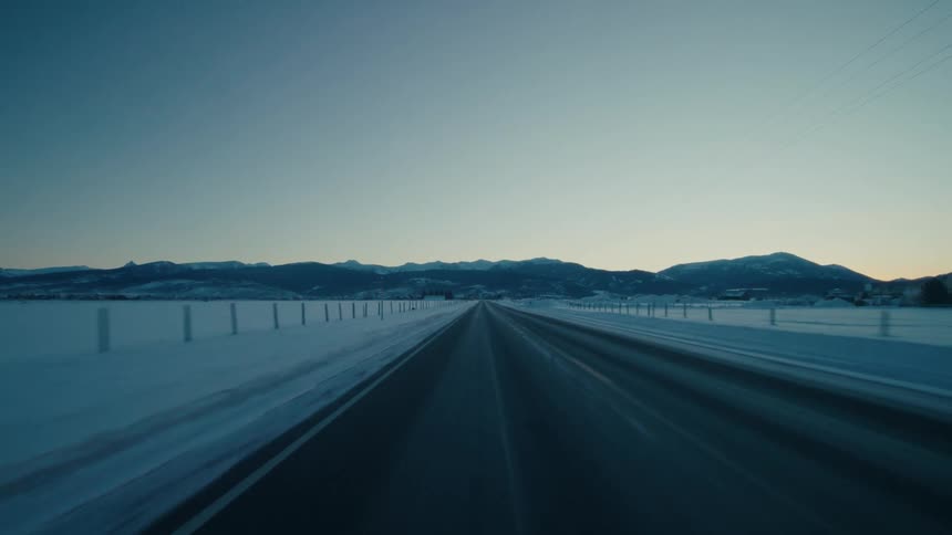 View of a the mountains on the horizon and of a road from the back of a moving vehicle riding through a frozen landscape