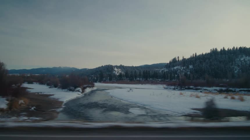 Traveling on a rural highway with snow and trees along the roadside and a stream with forested mountains in the distance