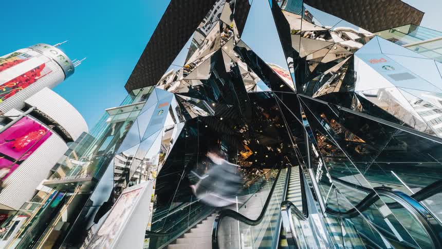 People enter building escalators as building mirrored surfaces reflect them