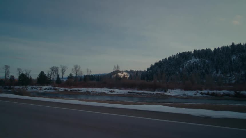 Traveling on a rural highway with snow and trees along the roadside and a stream with forested mountains in the distance