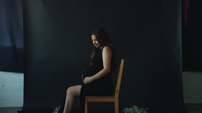 A pregnant woman sitting in a chair rubs her stomach before smiling into the camera
