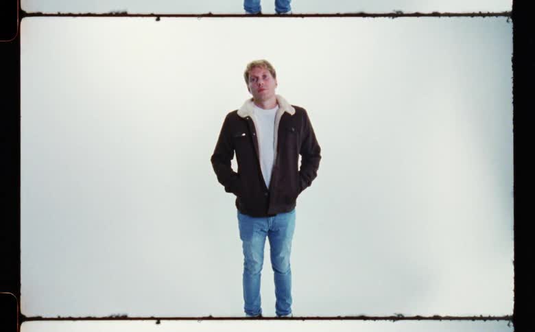 A man standing wearing a dark jacket and jeans with his hands in his coat pockets against a white backdrop