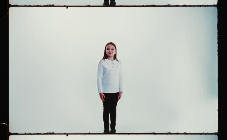 A little girl in a white sweater and black pants standing with her arms at her side against a white backdrop