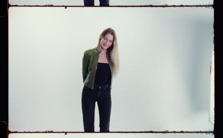Blonde woman with green jacket black top and pants poses for the camera in front of white screen backdrop in photography studio