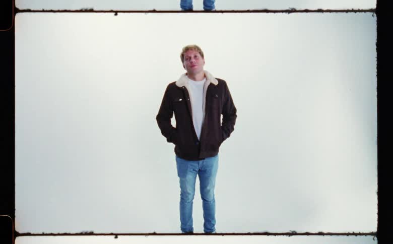 Man with black jacket and jeans stands with hands in jacket pockets in front of white screen backdrop in photography studio