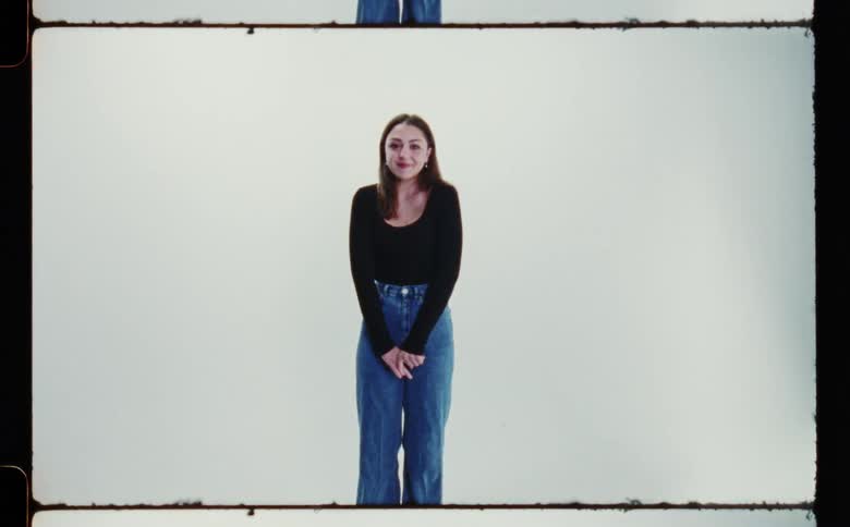 A teenage girl with long brown hair wearing a black shirt and blue jeans is looking ahead smiling and holding her hands together