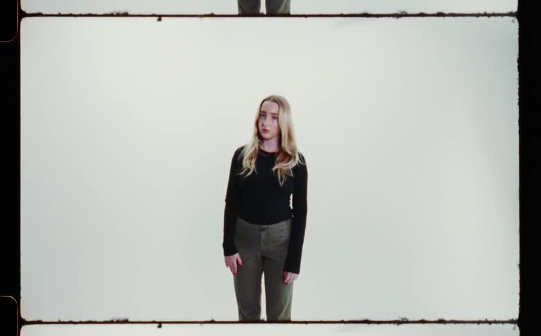 A teenage girl with long blond hair wearing a long sleeved black sweater and gray pants is standing and looking ahead