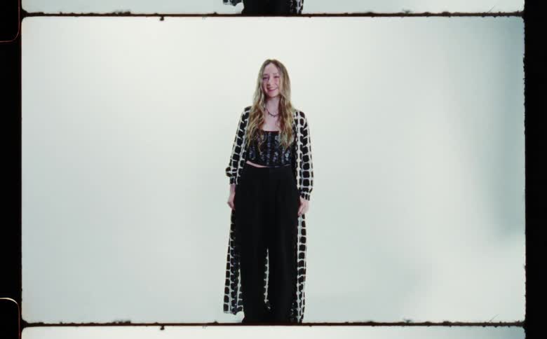 Blonde woman with white black overcoat black top and pants smiles at camera in front of white screen backdrop in photography studio
