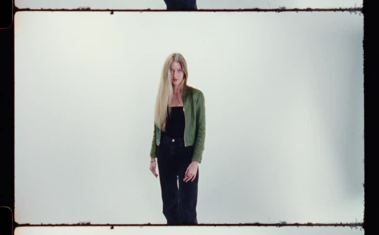 Blonde woman with green jacket black top and pants poses fort the camera in front of white screen backdrop in photography studio